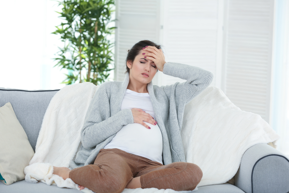Pregnant woman sitting on a sofa with a hand on her tummy and the other hand on her head. Signs of a headache